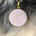 Pet ID tag hanging on a collar worn by a black and blonde dog. Made of gold plated brass and pink enamel, it reads 'Adopt Don't Shop'.