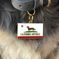 Pet ID tag hanging on a collar worn by a black and blonde dog. Made of gold plated brass with red and white enamel in the style of a California state flag but with a small dog instead of a bear.
