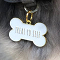 Pet ID tag hanging on a collar worn by a brown and white dog. It is made of gold plated brass and white enamel that is shaped like a bone and reads 'Treat Yo Self'.